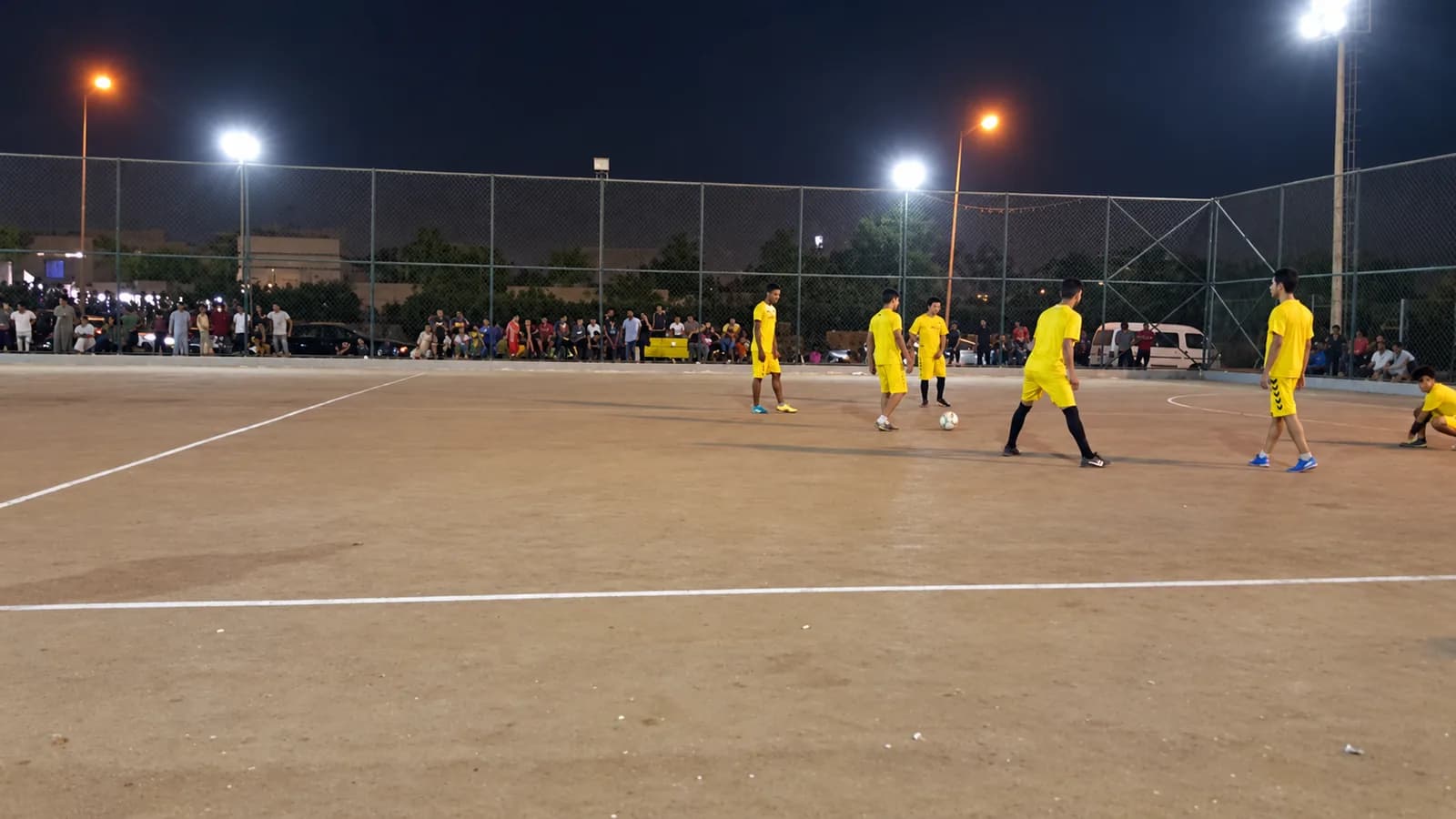 A five-a-side match under floodlights on the red clay pitch, players in yellow and white, spectators and parked cars along the fence