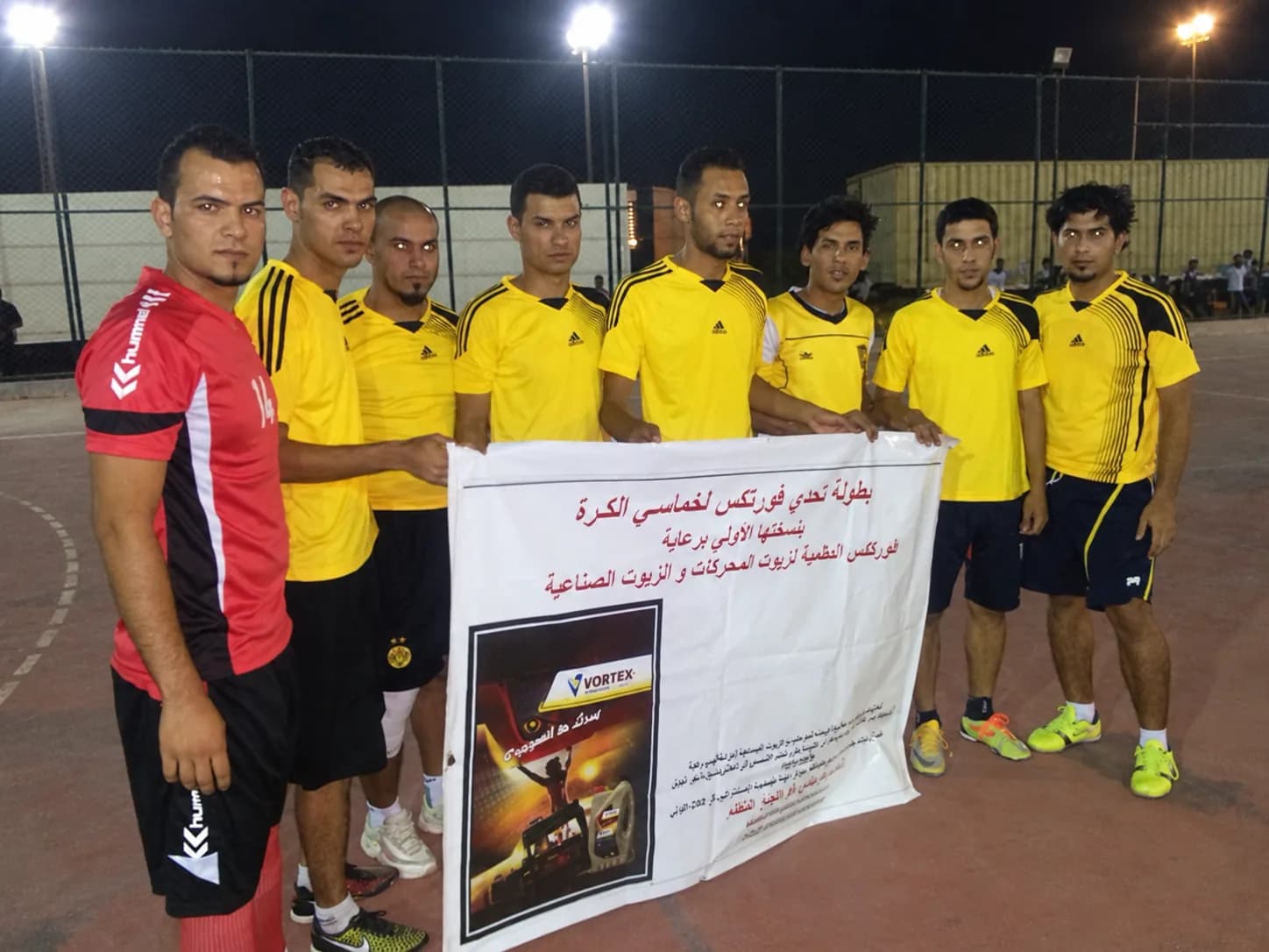 A team in yellow with the tournament banner, floodlights and spectators behind them